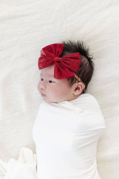 Newborn baby swaddled in white with a red bow headband on a white background