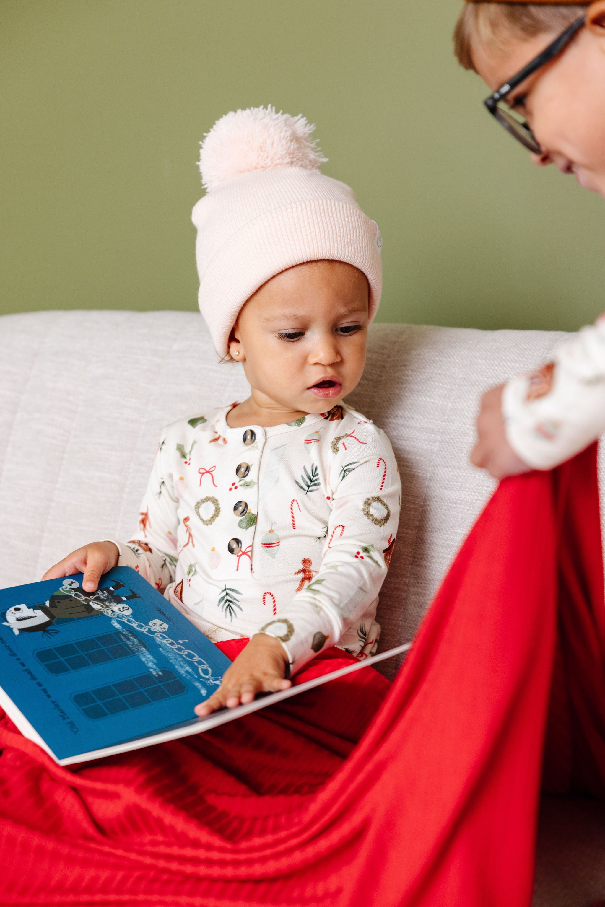 A toddler wearing a white knit pom-pom beanie and a white holiday print pajama set sits on a white couch, looking at a book while wrapped in a red blanket. Another person is partially visible on the right.