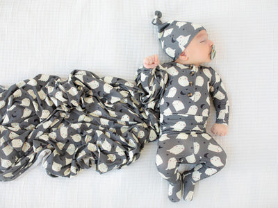 Baby in a gray and white patterned onesie and hat lying on a white blanket.