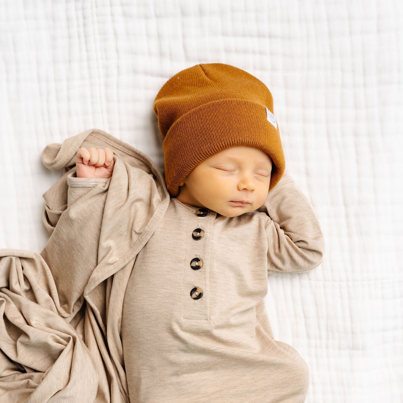 Newborn baby wrapped in a beige swaddle with a mustard yellow hat on a white textured background