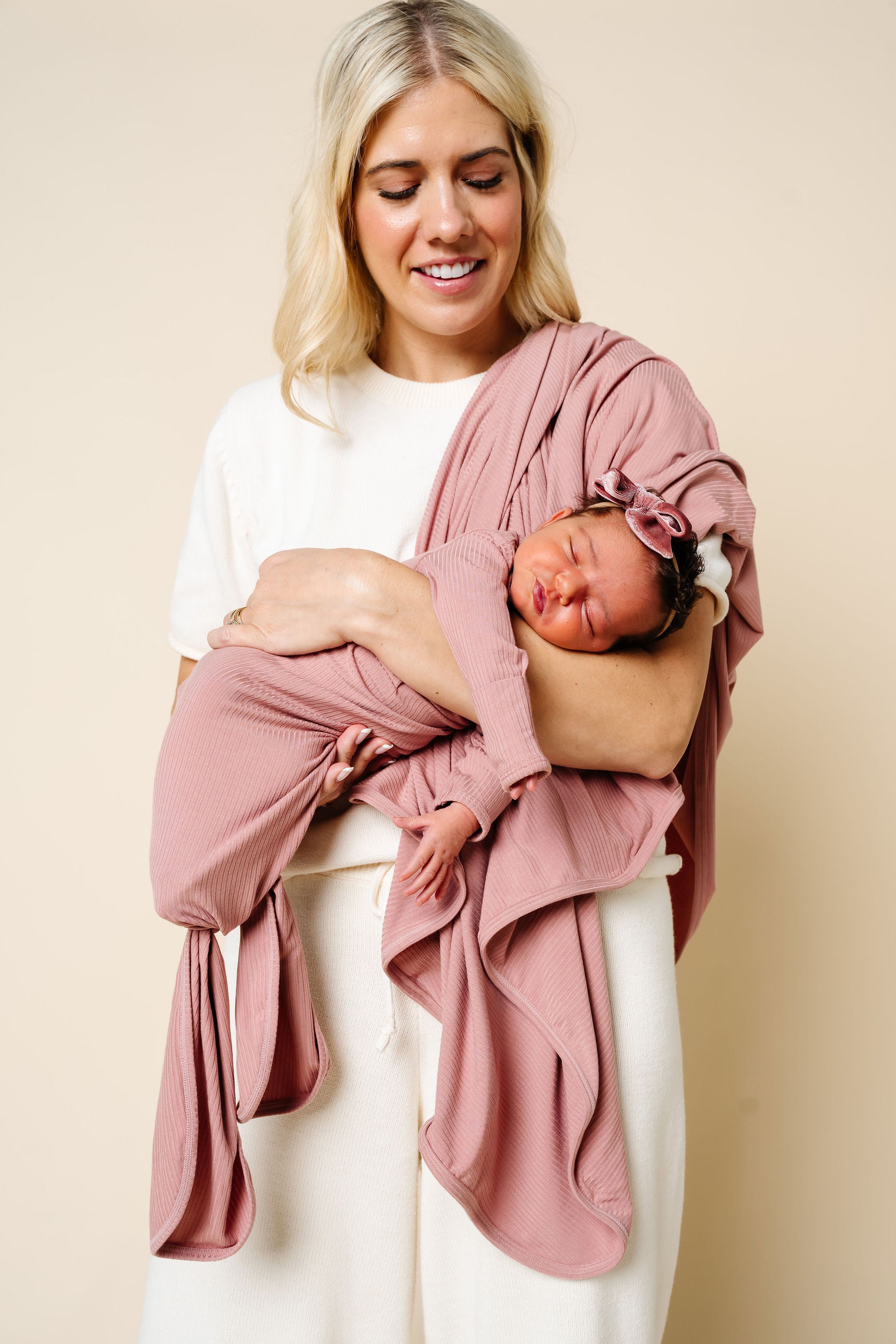 Woman holding a baby wrapped in a pink wrap against a beige background
