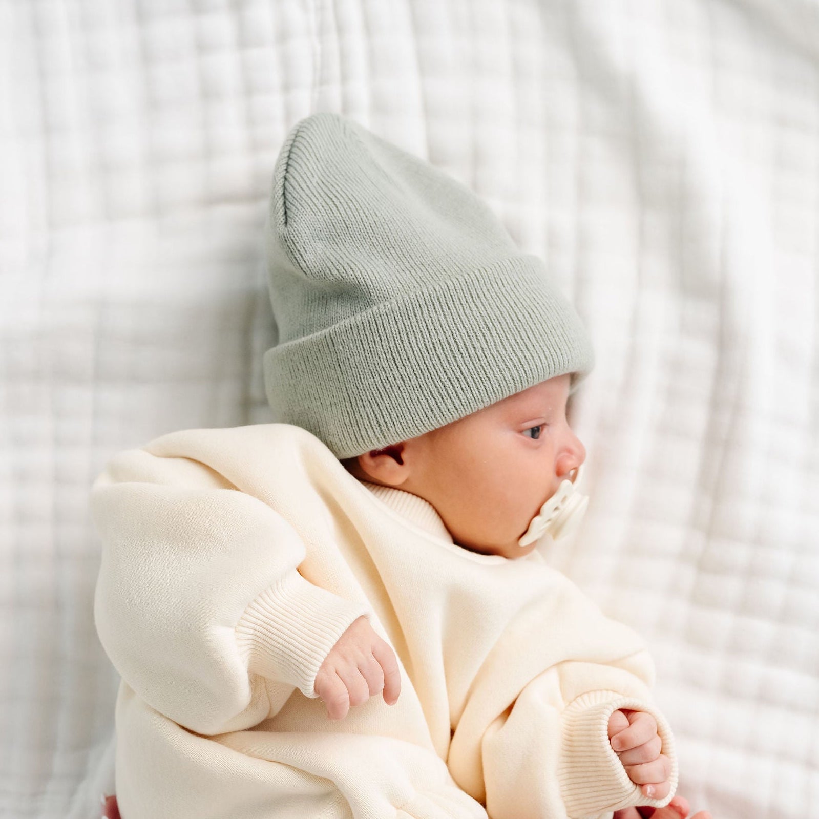 Newborn baby in a white outfit and gray hat held by a person on a white blanket.