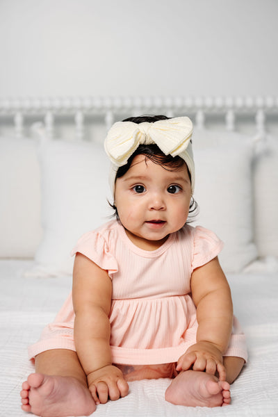 Baby wearing a pink dress and white headband with a bow, sitting on a white surface.