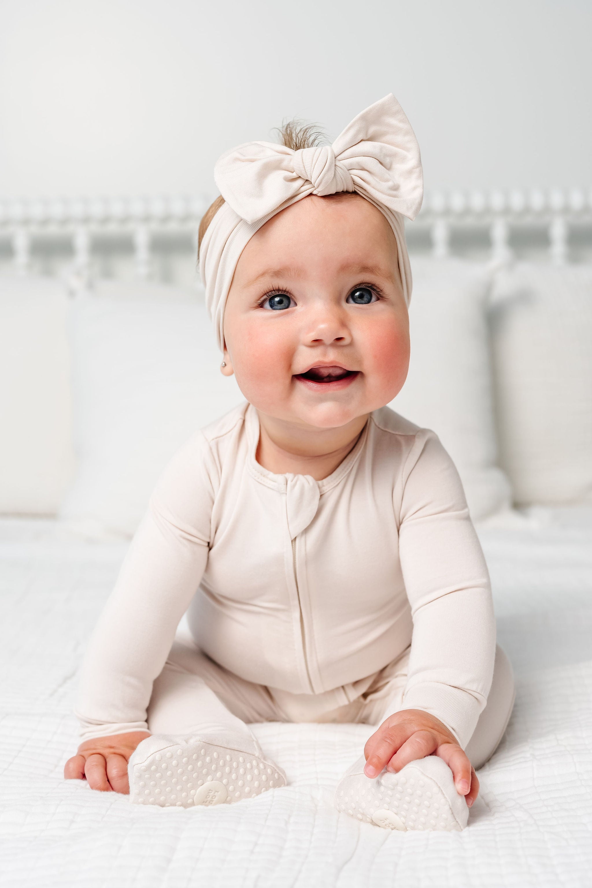Baby wearing a beige outfit and headband sitting on a white bed.