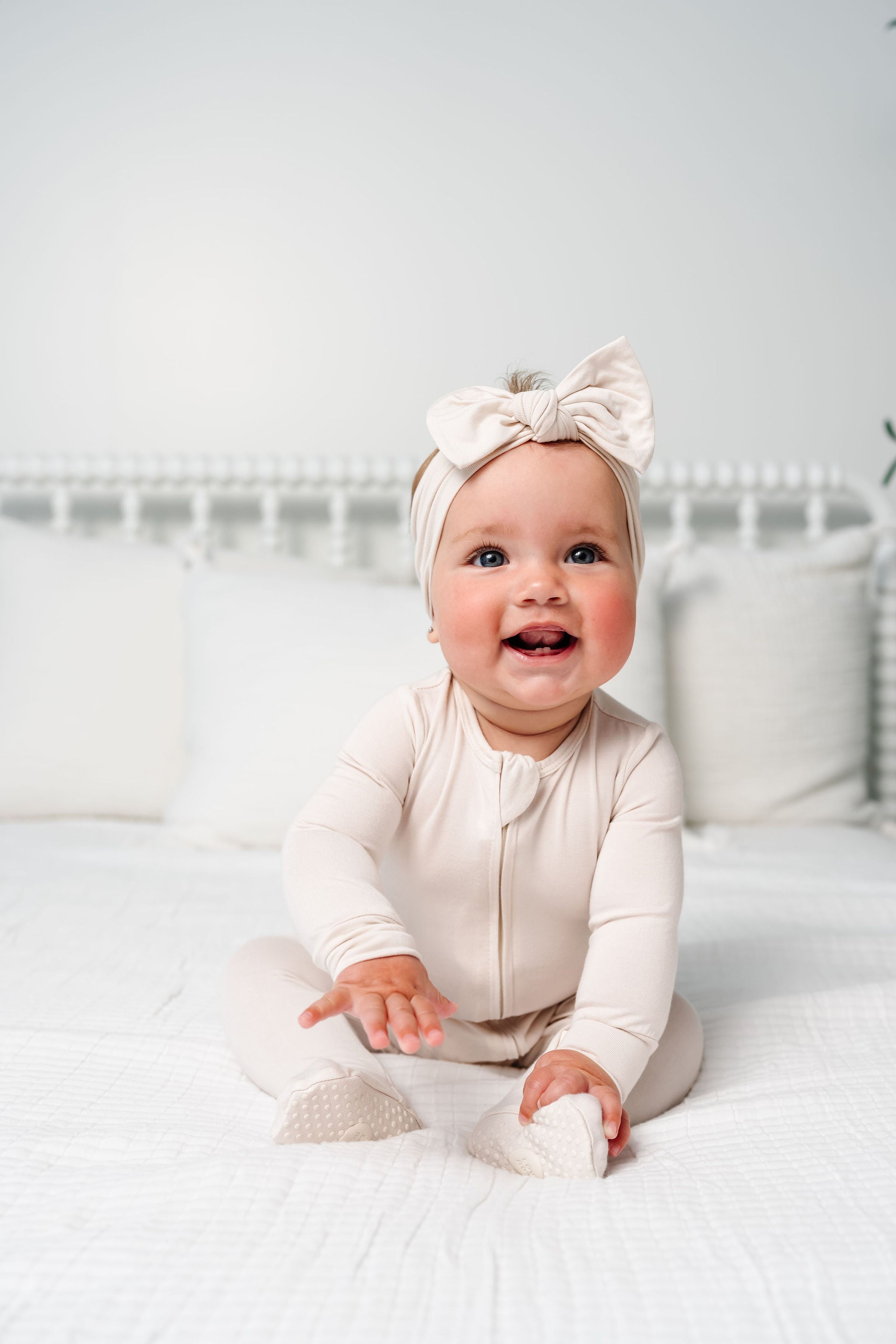 Baby in a white outfit with a bow headband sitting on a bed.