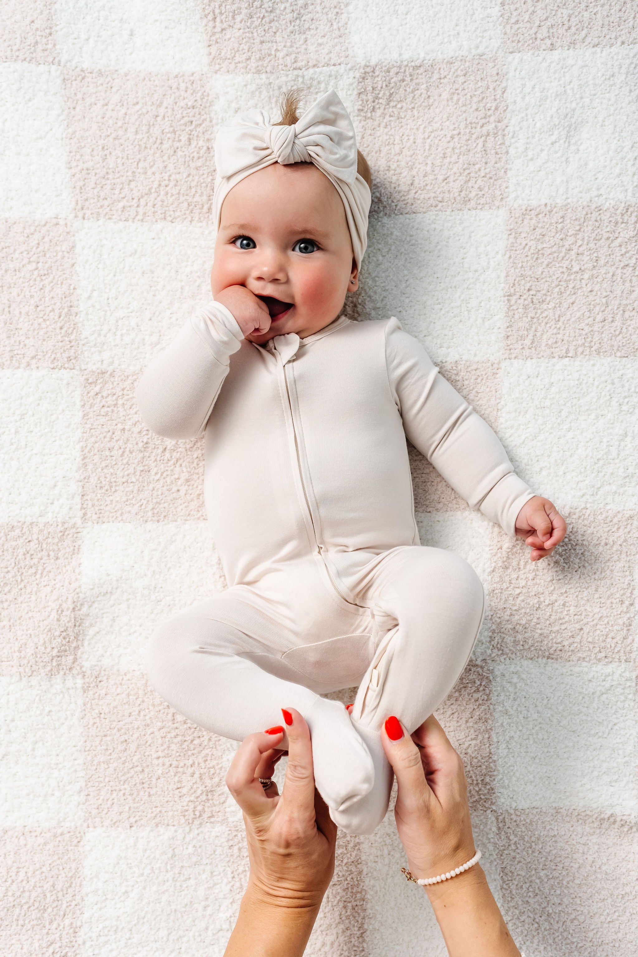 Baby in a white outfit lying on a textured surface with a person's hands holding its feet.
