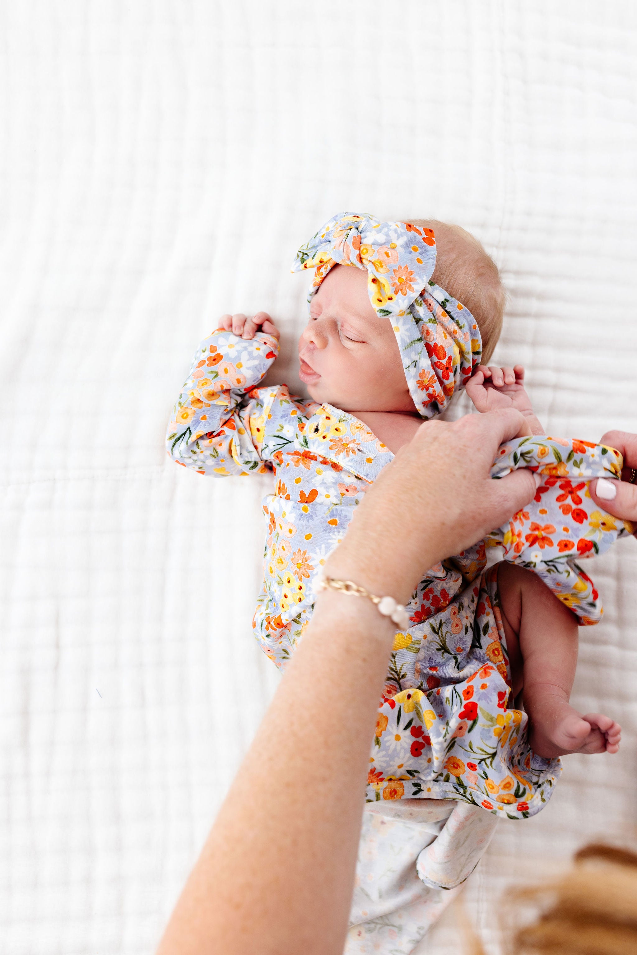 Newborn baby in floral outfit and headband being held on a white blanket