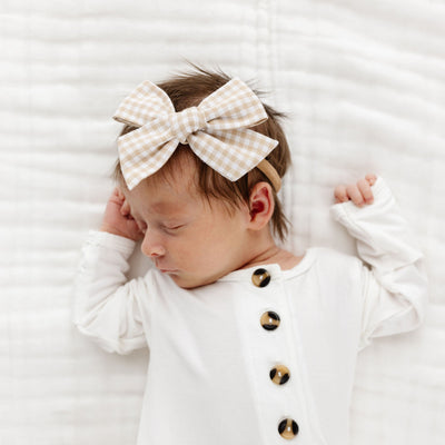 Baby wearing a white outfit with gold buttons and a large bow headband, lying on a white textured surface.