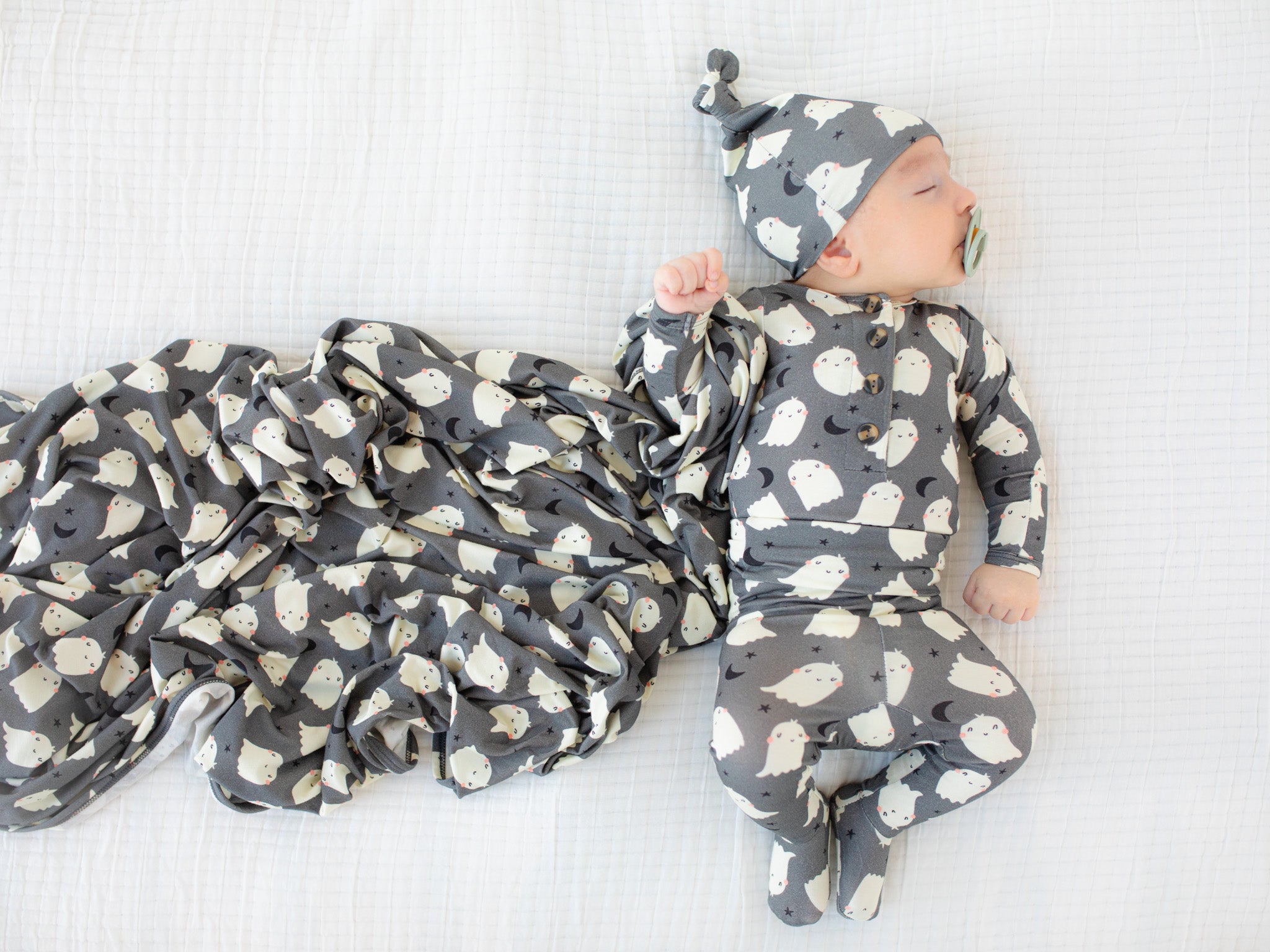 Baby in a gray and white patterned onesie and hat lying on a white blanket.