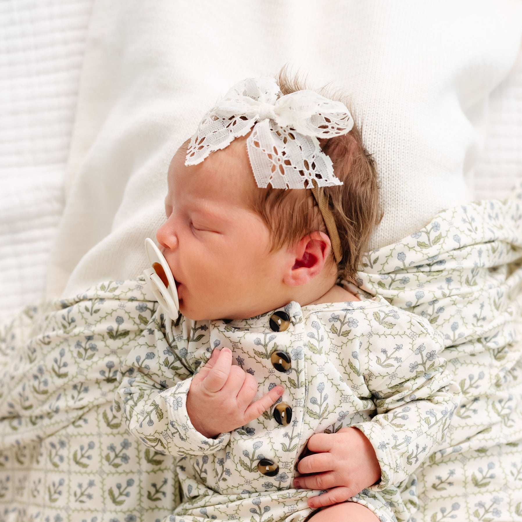 Newborn baby in a floral outfit with a bow headband, lying on a white blanket.