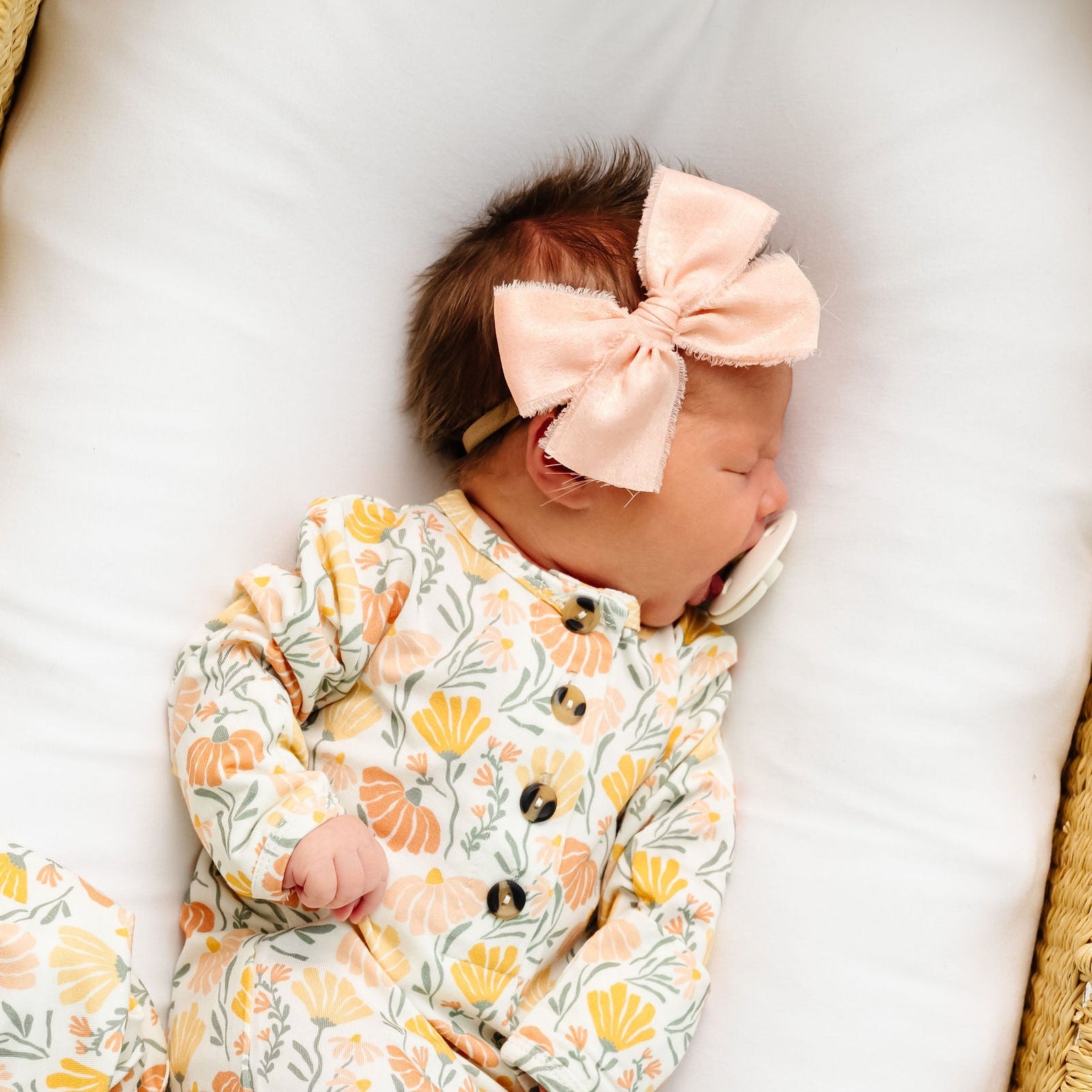 Newborn baby in a floral outfit with a pink bow in a wicker crib.