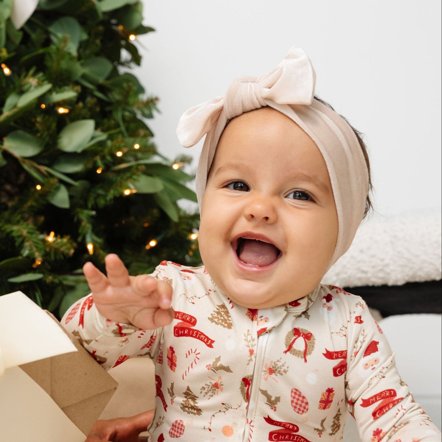 Baby in festive pajamas holding a gift box with a Christmas tree in the background
