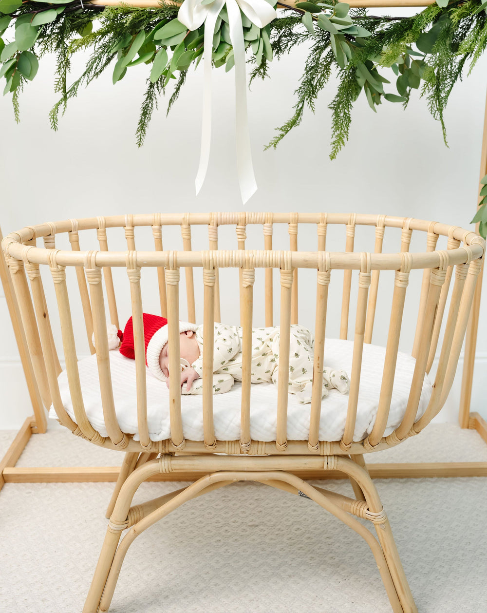 Wooden crib with baby wearing a red hat and white bear plush toy, decorated with greenery and a white bow.