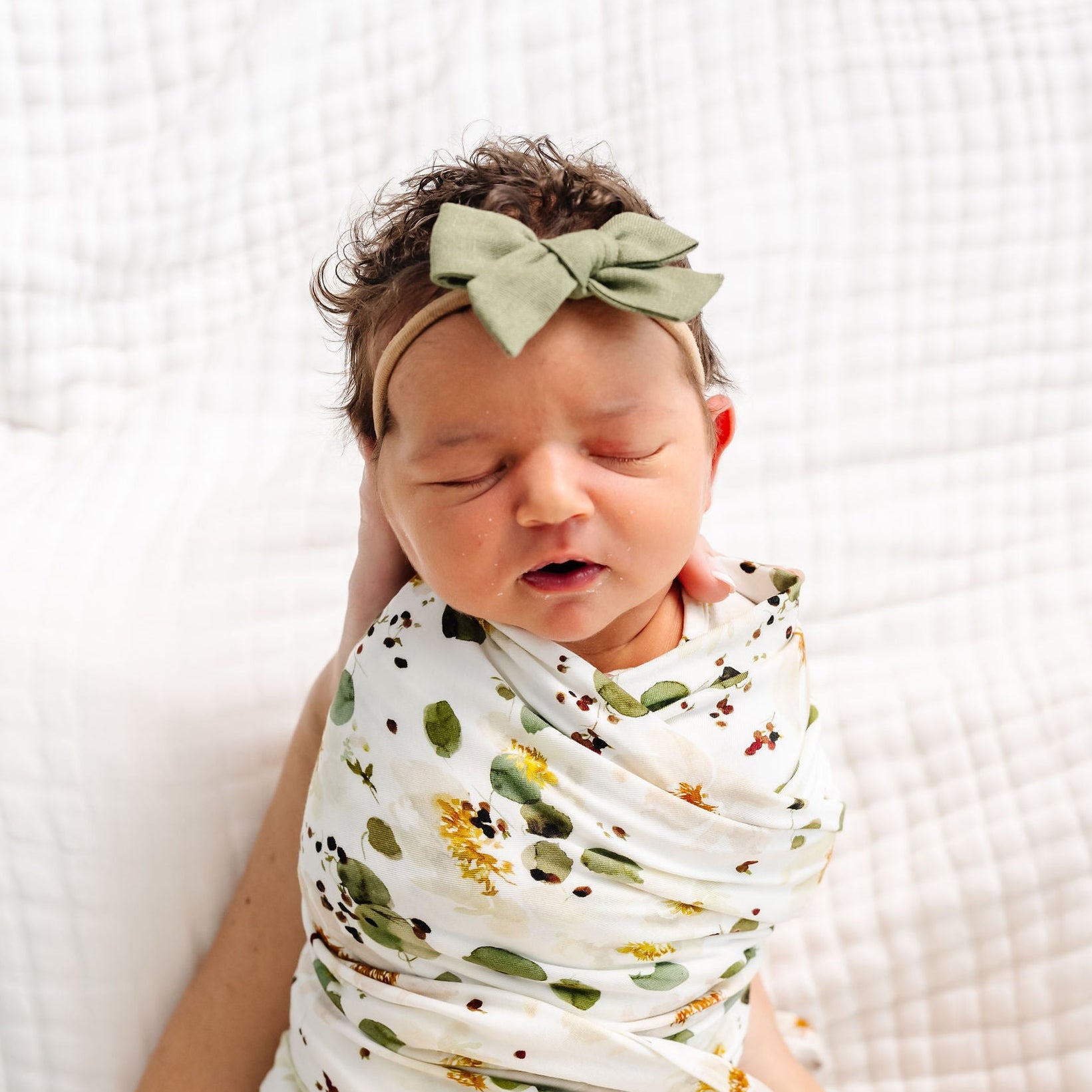 Newborn baby swaddled in a floral blanket with a green headband, sitting on a white textured surface.