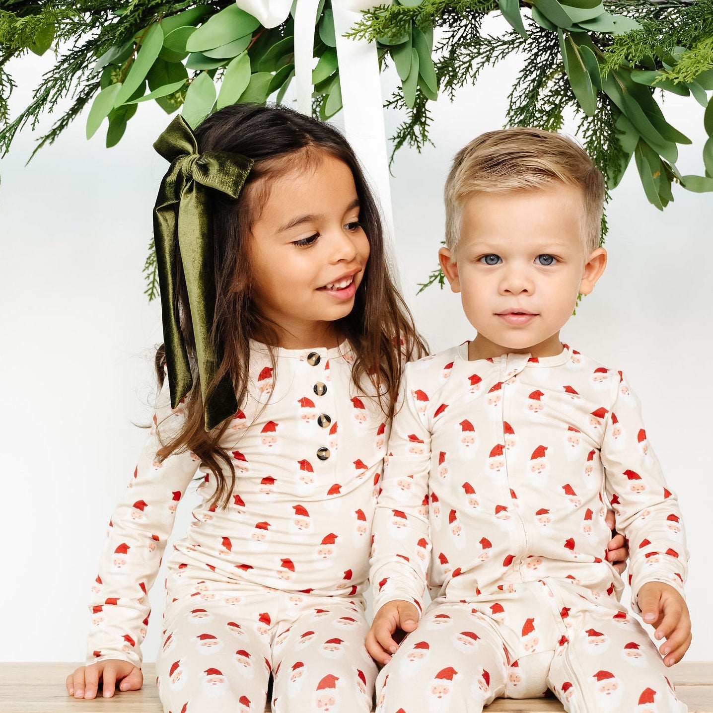 Two children in matching pajamas sitting on a wooden bench with greenery above.