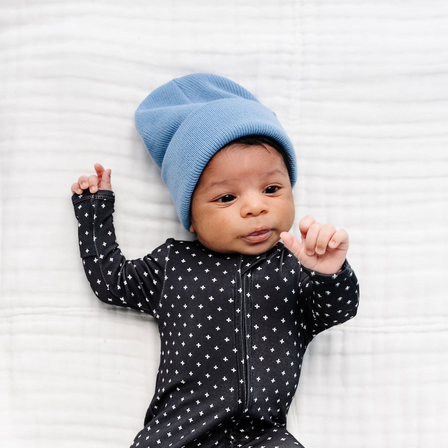 Baby wearing a blue hat and black polka dot outfit lying on a white textured surface