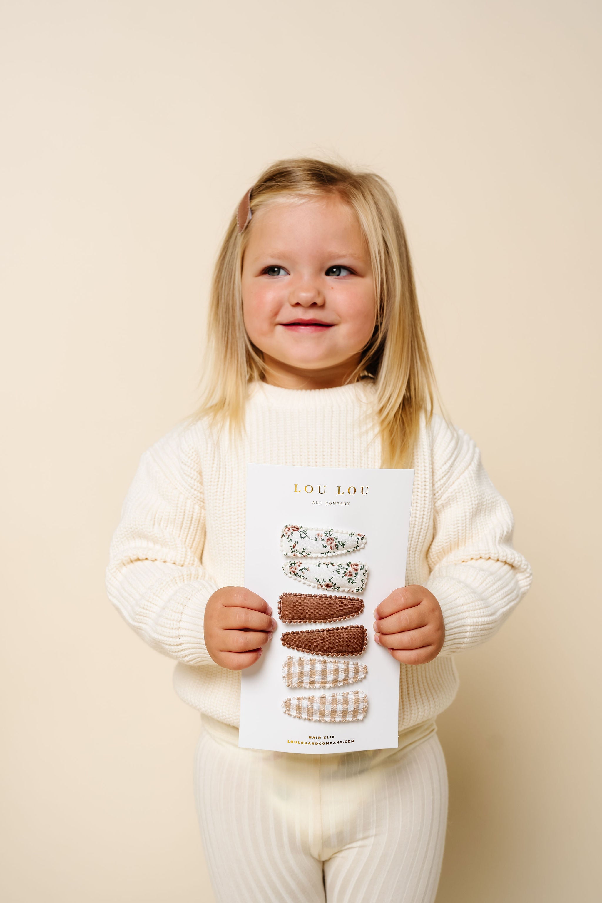 Child holding a product packaging with 'Lou Lou' branding against a beige background