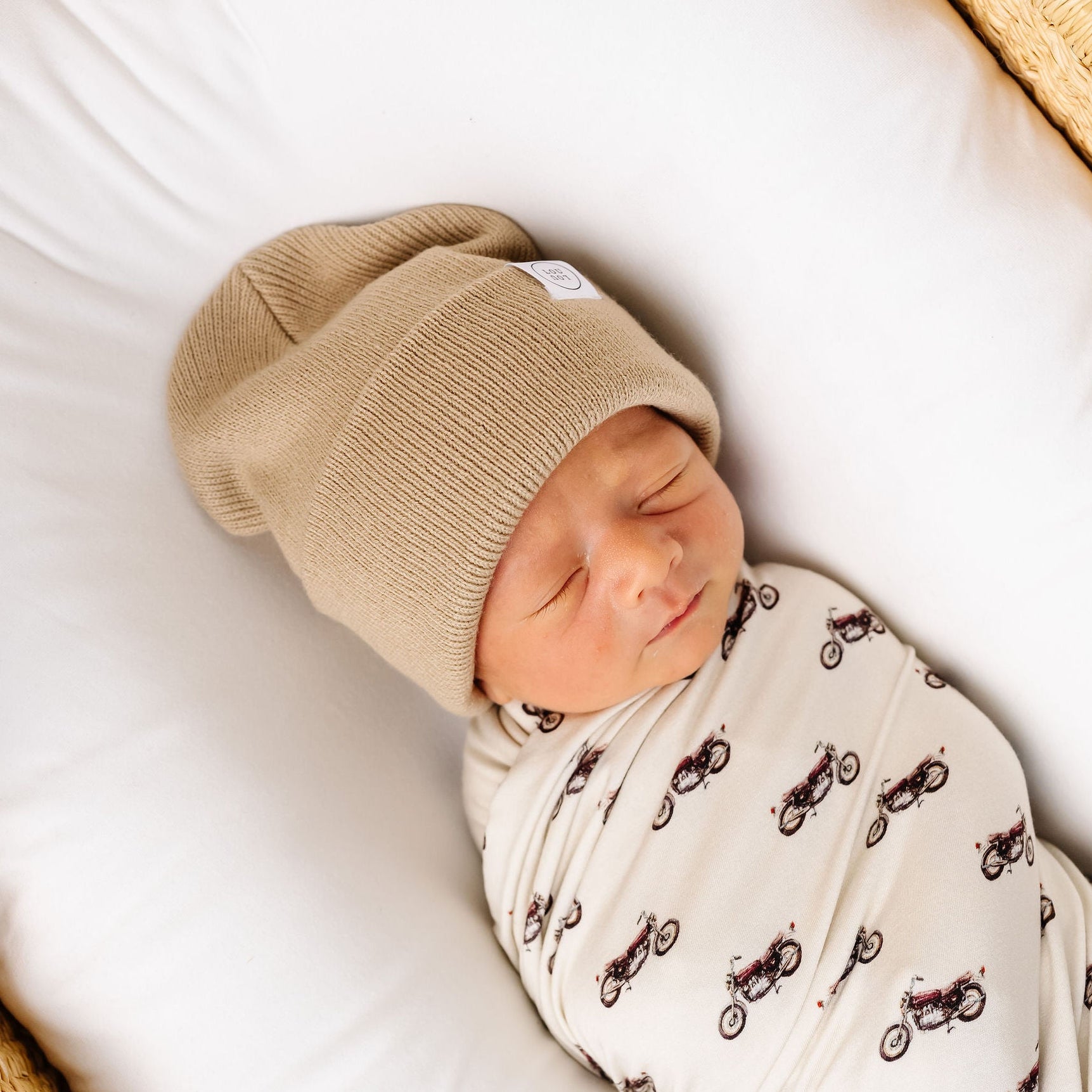 Newborn baby swaddled in a blanket with a pattern, wearing a beige hat, in a crib.